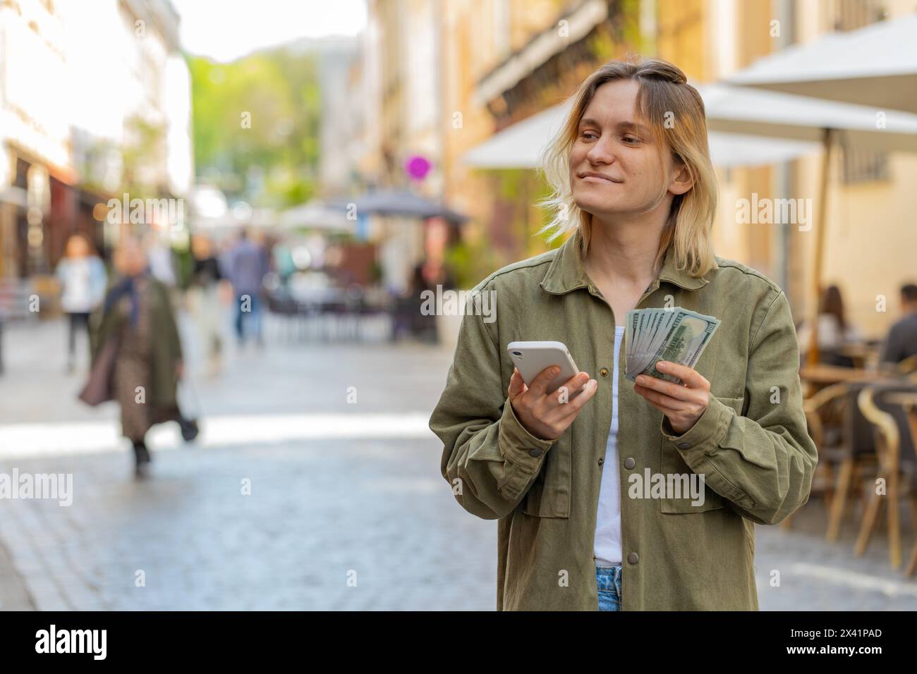 Heureuse jeune femme comptant argent dollar cash, en utilisant l'application de calculatrice de smartphone, satisfaite du revenu pour les cadeaux de vacances prévus. Fille caucasienne sur la rue ensoleillée de la ville urbaine. Mode de vie urbain en plein air. Banque D'Images