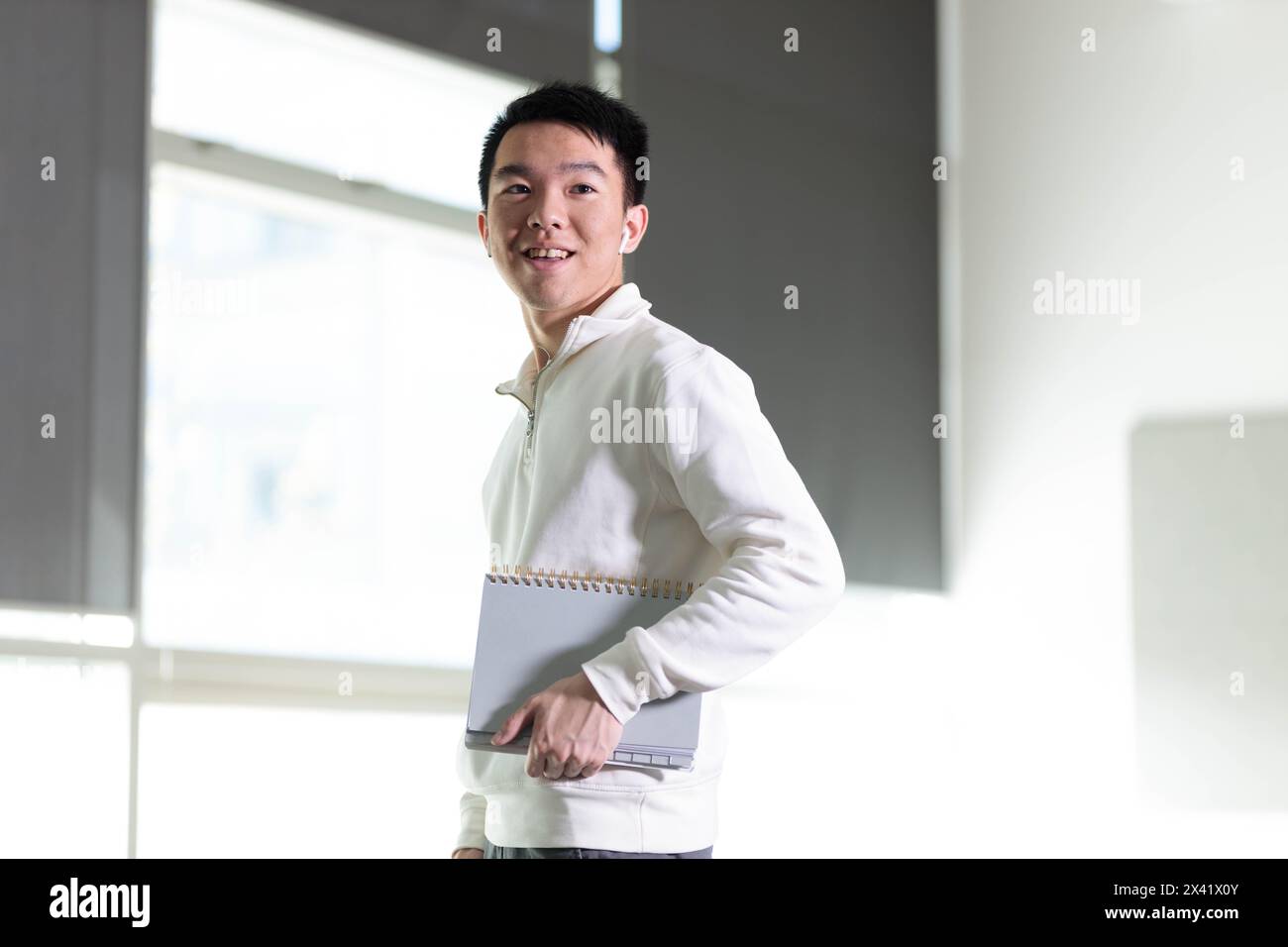 Un étudiant en pull blanc sourit tout en tenant un livre et un téléphone portable. Il est debout dans une salle de classe d'université ou de lycée tout en écoutant Banque D'Images