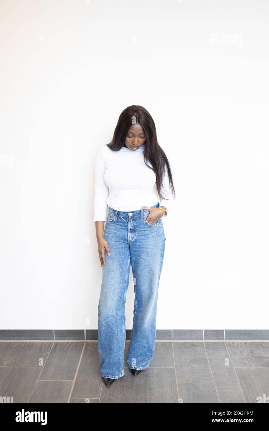 Une jeune femme noire prend une pose détendue contre un mur blanc minimaliste. Ses cheveux longs et lisses complètent l'élégance intemporelle d'un col roulé blanc ajusté, parfaitement associé à un Jean coupe large chic, créant un équilibre entre style classique et contemporain. Femme élégante en Jean blanc à col roulé et coupe large. Photo de haute qualité Banque D'Images