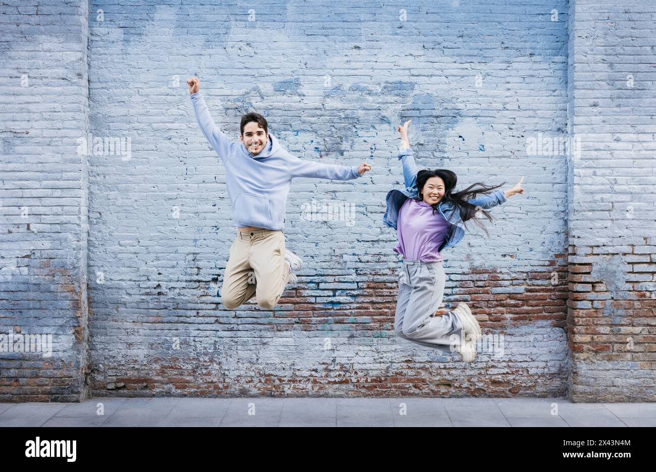 Jeune homme et femme sautant par le mur de briques bleues. Couple sautant joyeusement contre un mur coloré et souriant. Banque D'Images