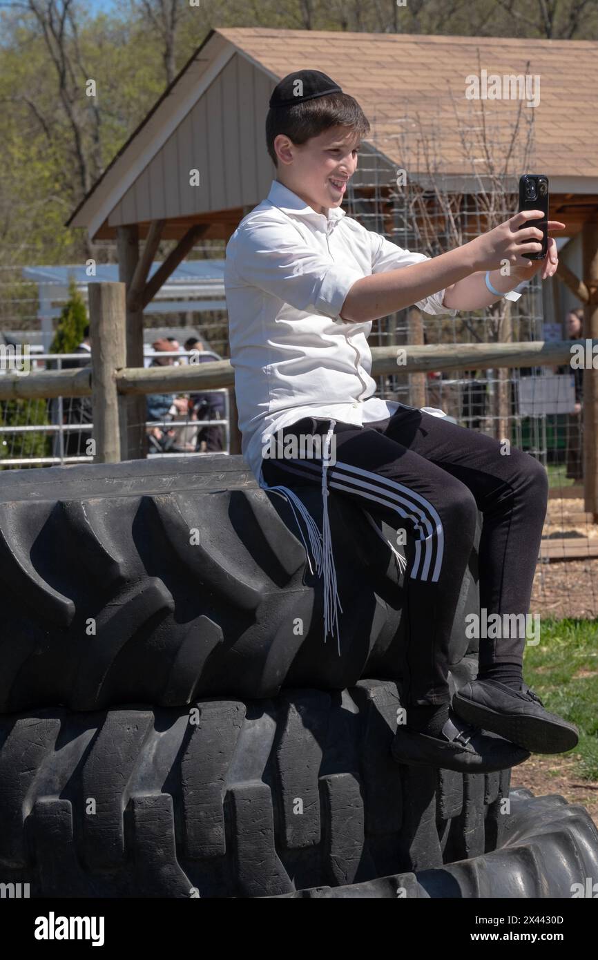 Un adolescent juif orthodoxe prend un selfie assis sur une pile de pneus surdimensionnés. Dans une ferme du comté de Rockland et pendant la Pâque 2024. Banque D'Images