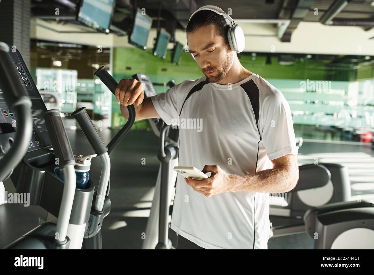 Un homme concentré en sport portant des écouteurs et utilisant un téléphone portable tout en s'entraînant dans une salle de gym. Banque D'Images