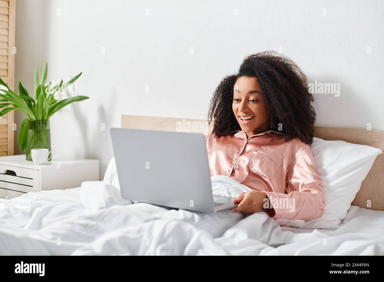 Femme afro-américaine en pyjama assis sur le lit, en utilisant un ordinateur portable dans la chambre le matin. Banque D'Images