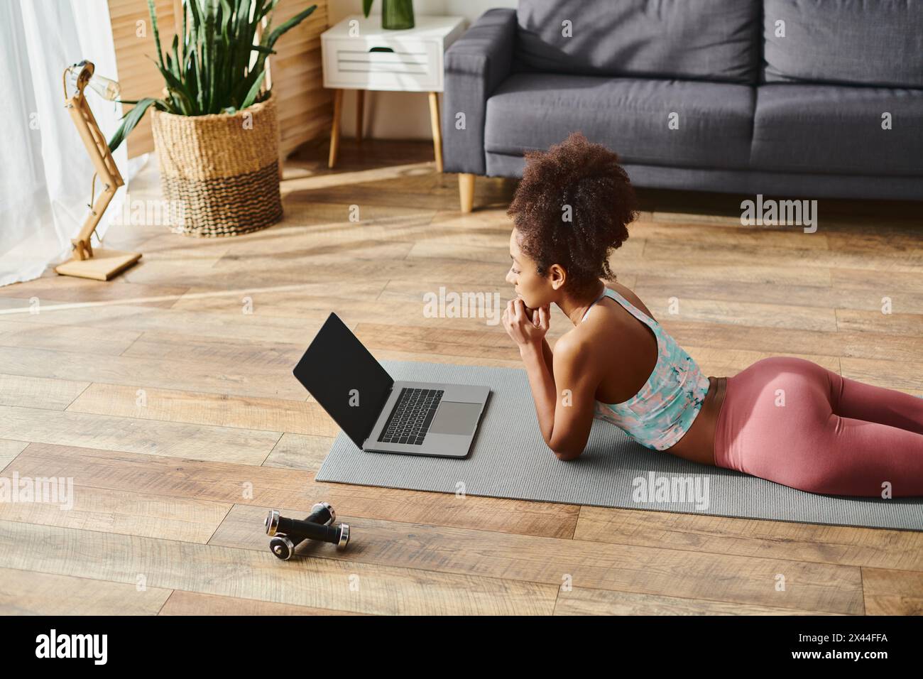 Femme afro-américaine bouclée dans des vêtements d'activité assis sur un tapis de yoga, en utilisant un ordinateur portable pour l'instruction de fitness virtuelle. Banque D'Images