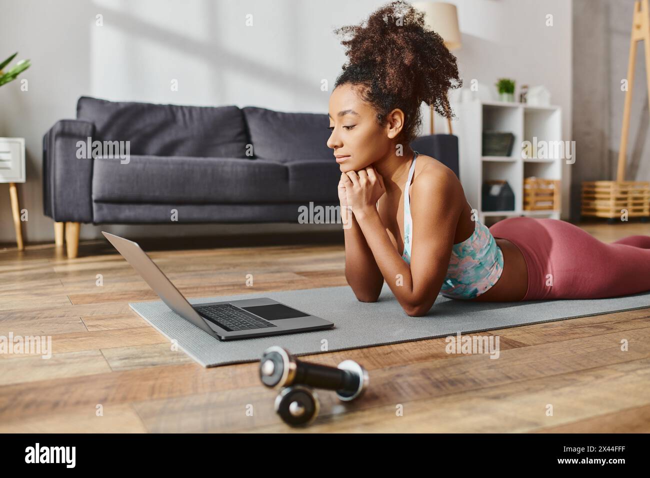 Une femme afro-américaine bouclée en vêtements sportifs pratique le yoga tout en utilisant un ordinateur portable sur un tapis à la maison. Banque D'Images