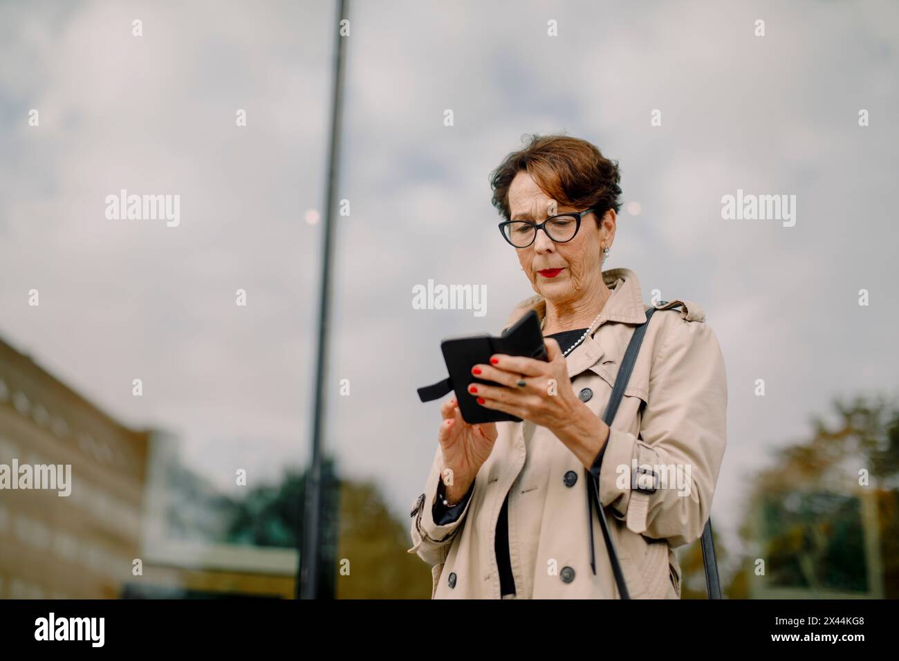 Femme âgée retraitée utilisant un téléphone intelligent contre un mur de verre Banque D'Images
