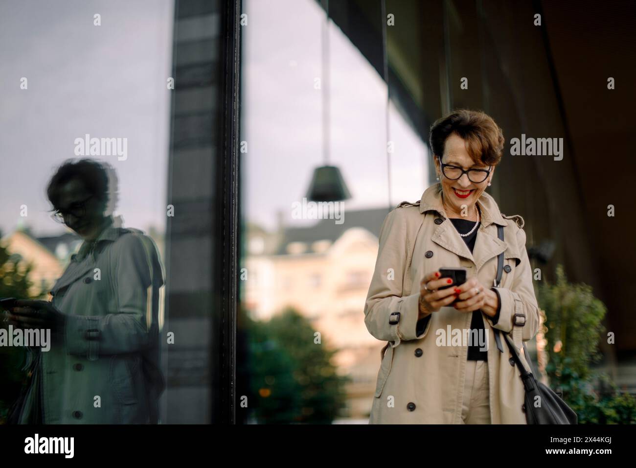 Femme âgée souriante à l'aide d'un téléphone intelligent tout en se tenant par la réflexion de verre Banque D'Images