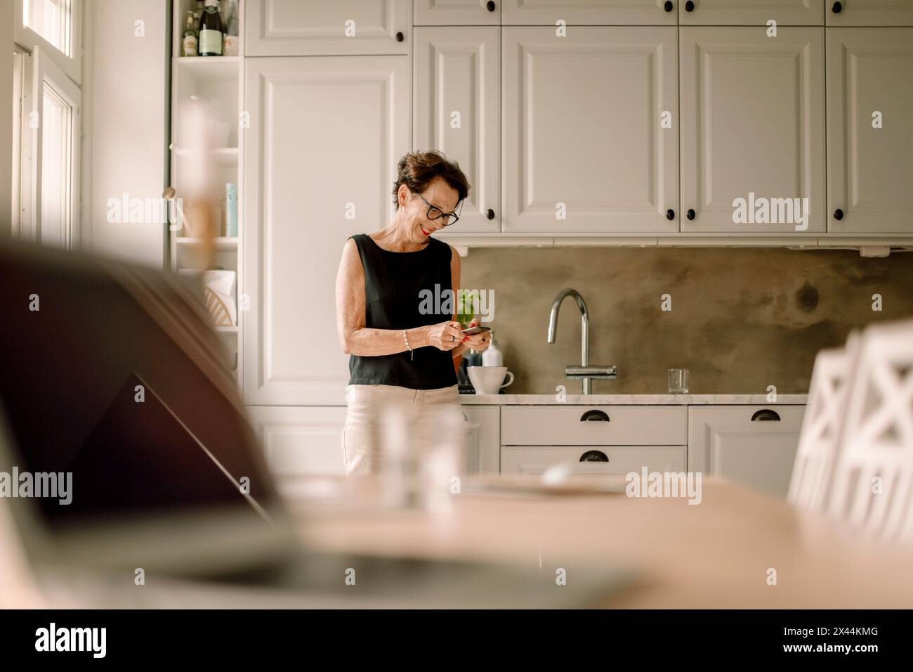 Femme senior retraitée utilisant un téléphone intelligent tout en s'appuyant sur le comptoir de cuisine Banque D'Images