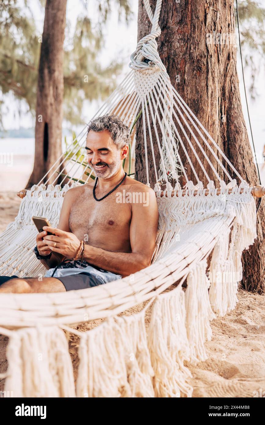 Sourire homme torse à l'aide d'un téléphone intelligent tout en étant assis sur un hamac à la plage Banque D'Images