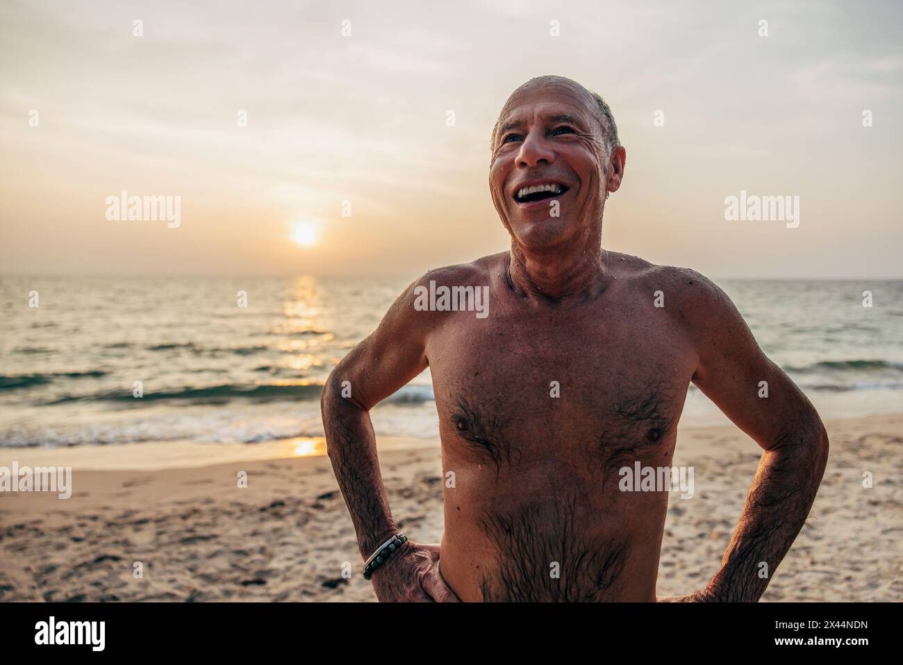 Homme senior sans torse insouciant debout avec les bras akimbo à la plage Banque D'Images