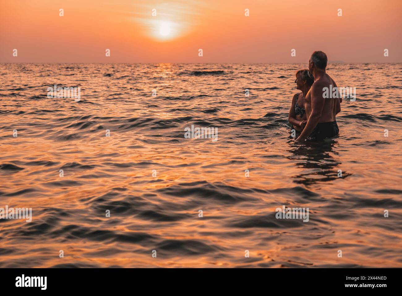 Couple senior insouciant regardant le coucher du soleil tout en se tenant dans la mer en vacances Banque D'Images