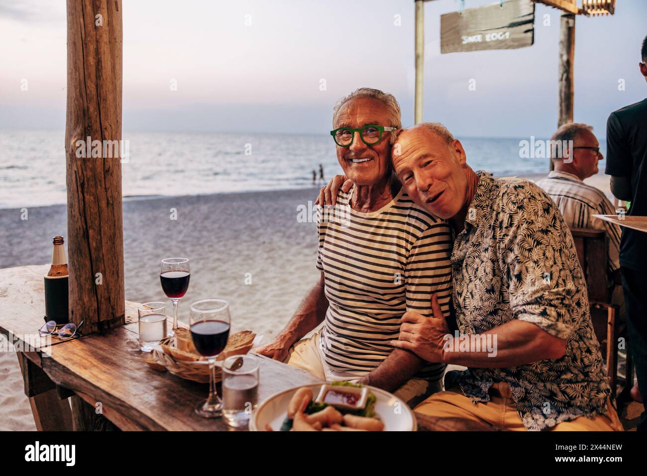 Portrait de couple gay heureux avec du vin et des collations sur la table au restaurant de plage Banque D'Images