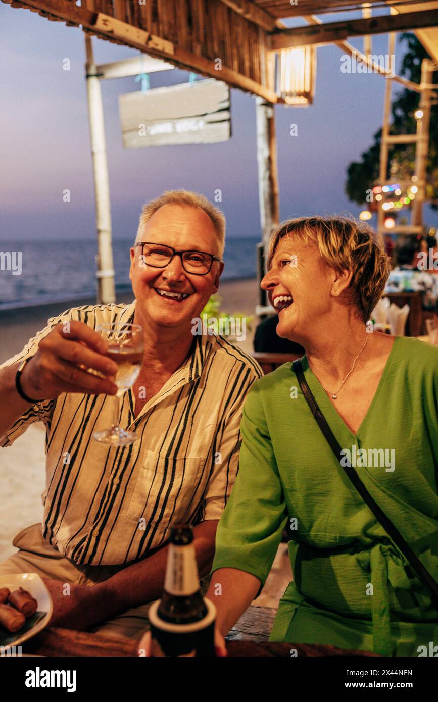 Portrait d'un homme senior souriant appréciant le vin avec une femme assise au restaurant de plage Banque D'Images