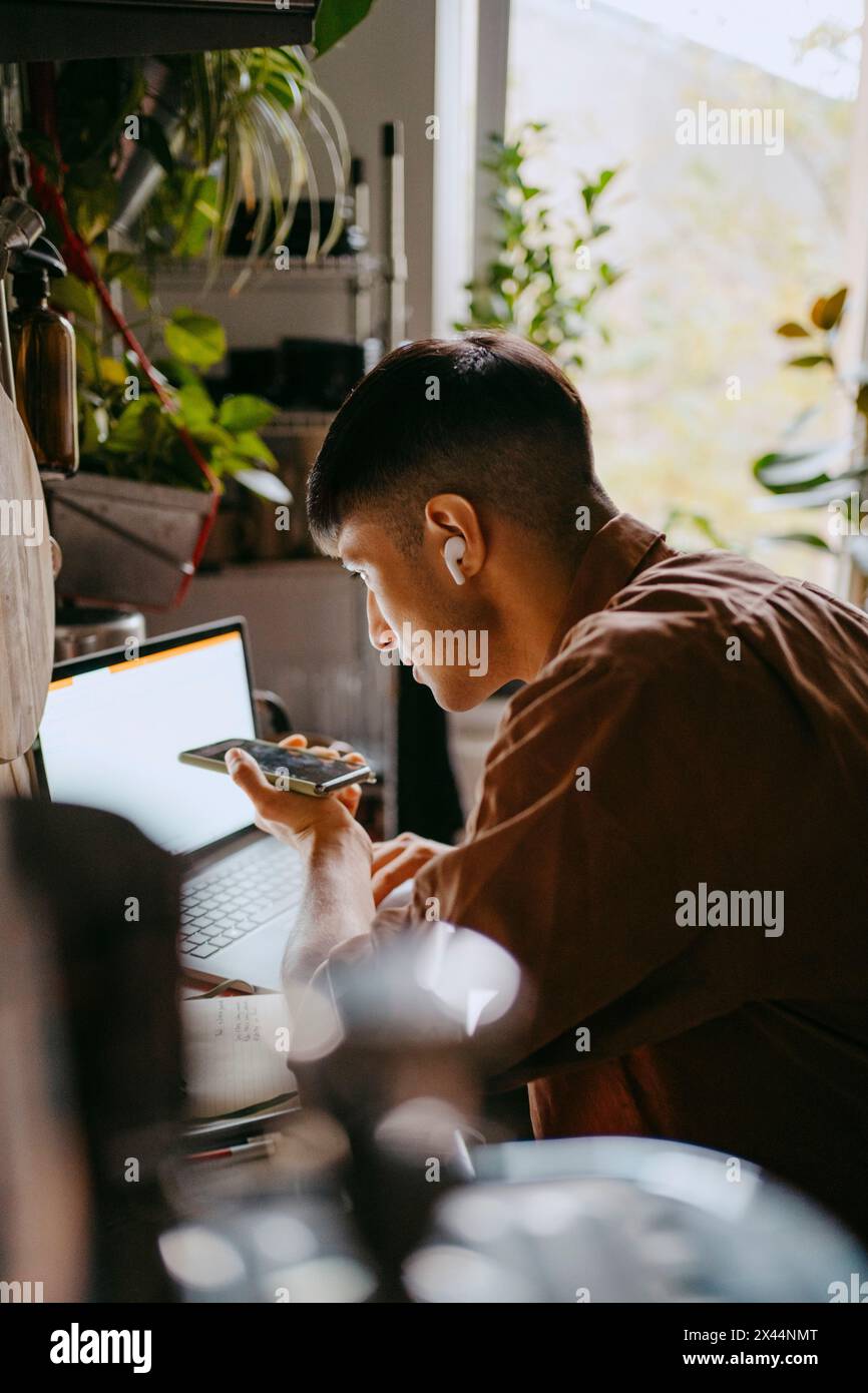 Homme freelance parlant sur le téléphone à haut-parleur tout en utilisant un ordinateur portable dans la cuisine au bureau à domicile Banque D'Images