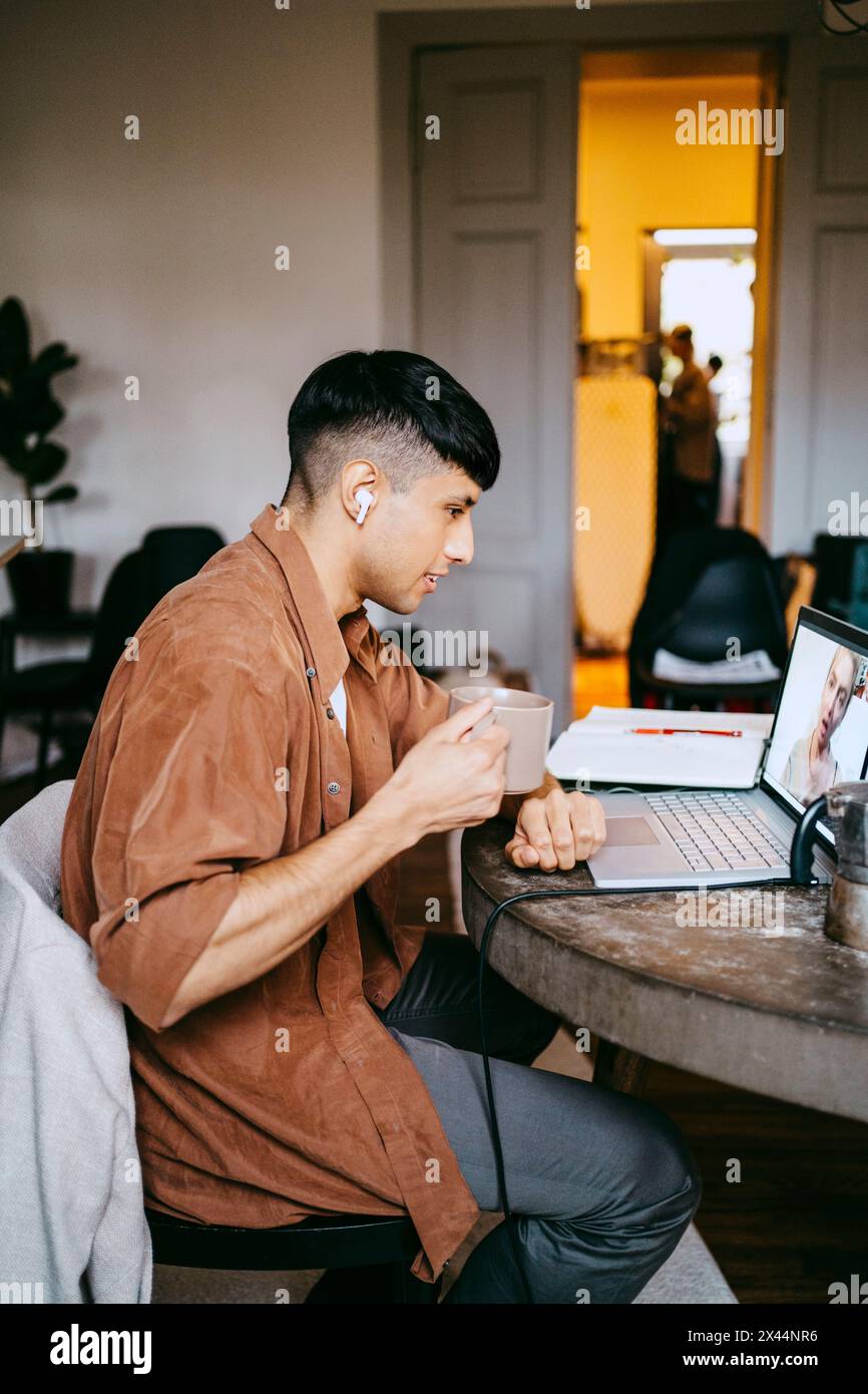 Vue latérale d'un freelance masculin prenant un café tout en faisant un appel vidéo avec un collègue sur ordinateur portable au bureau à domicile Banque D'Images