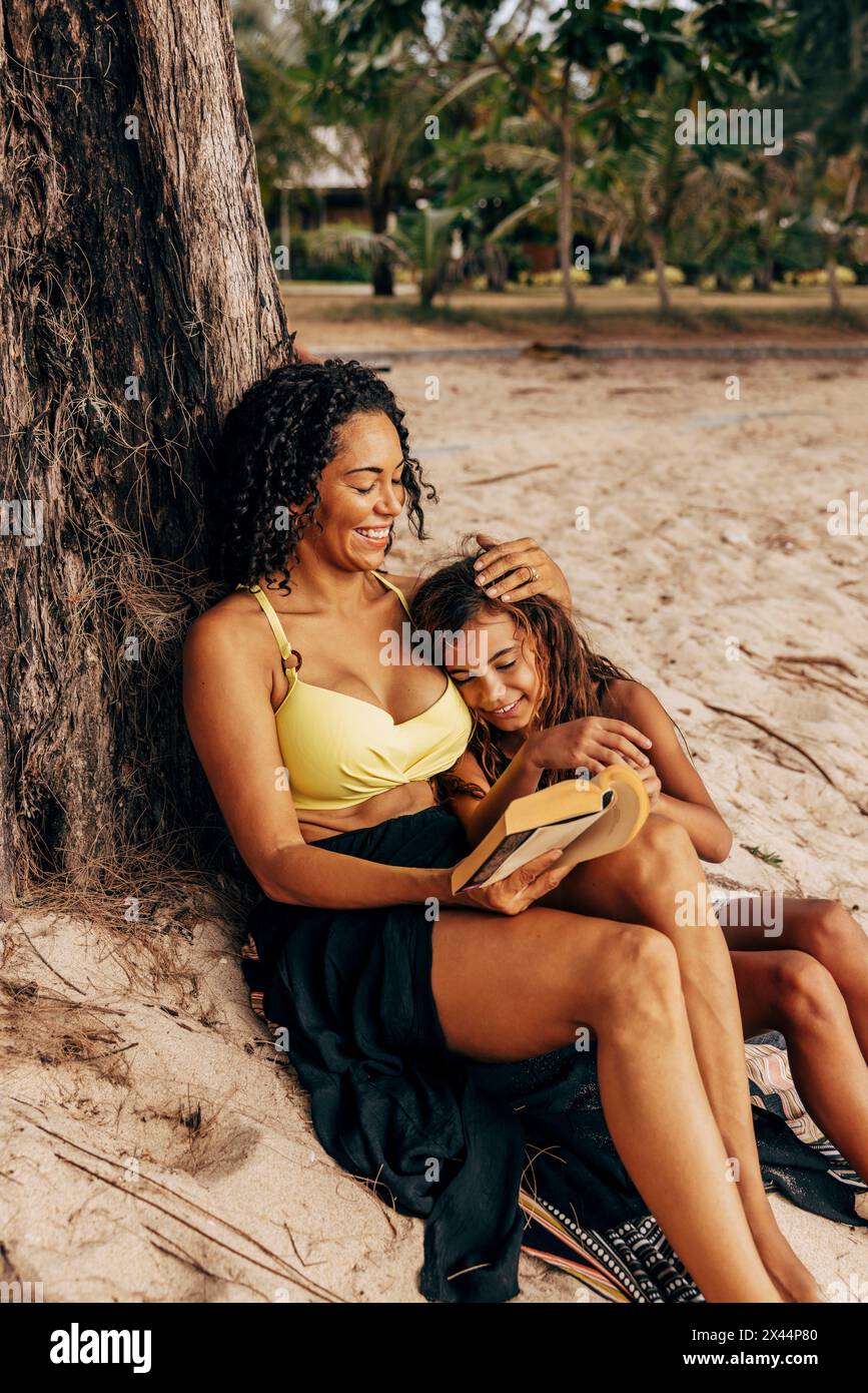 Mère souriante caressant sa fille lisant le livre tout en étant assis près de l'arbre à la plage Banque D'Images