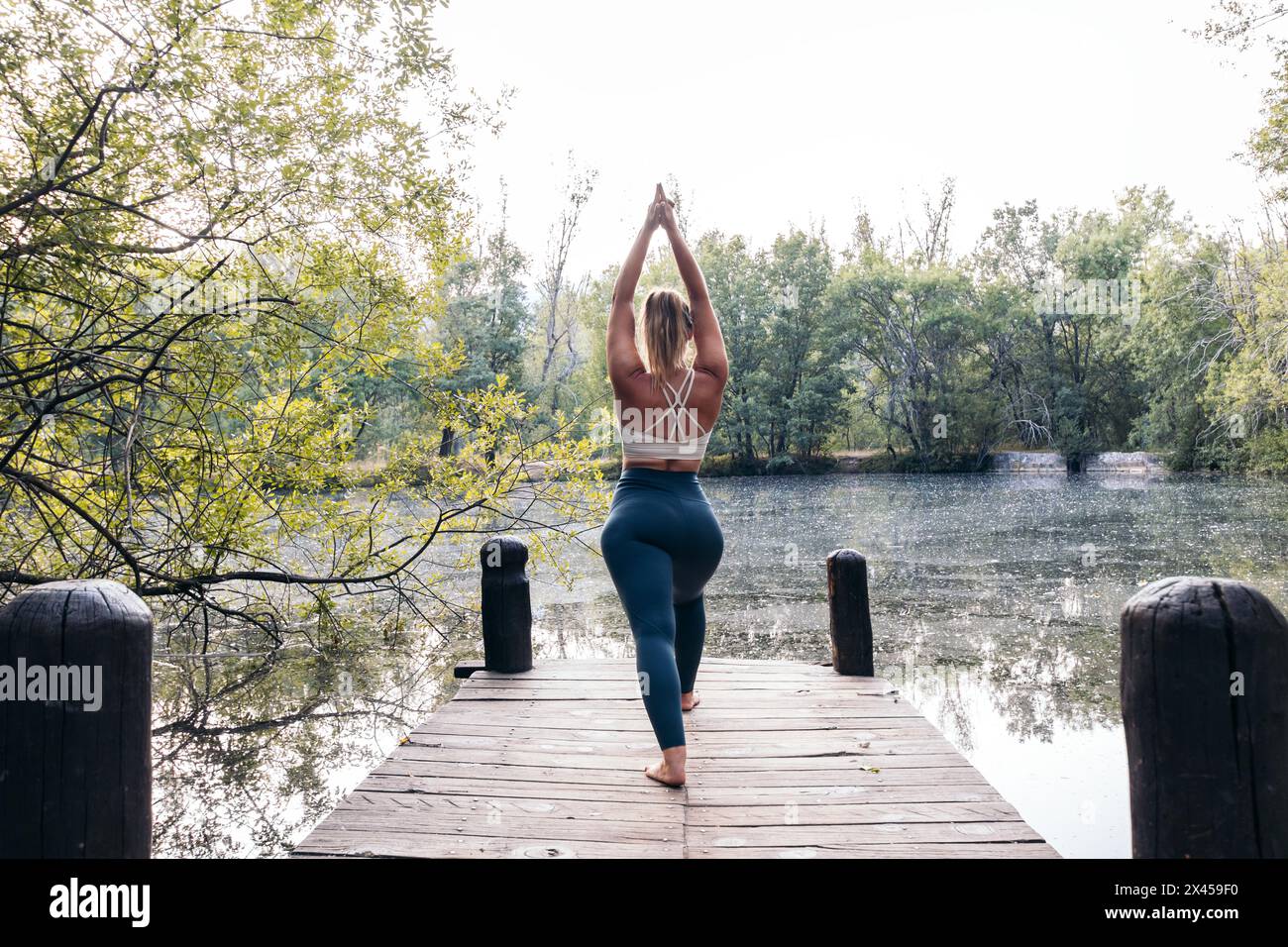Vue arrière d'une jeune femme athlétique faisant du yoga dans la nature. Elle est pieds nus sur une plate-forme en bois près d'un lac Banque D'Images