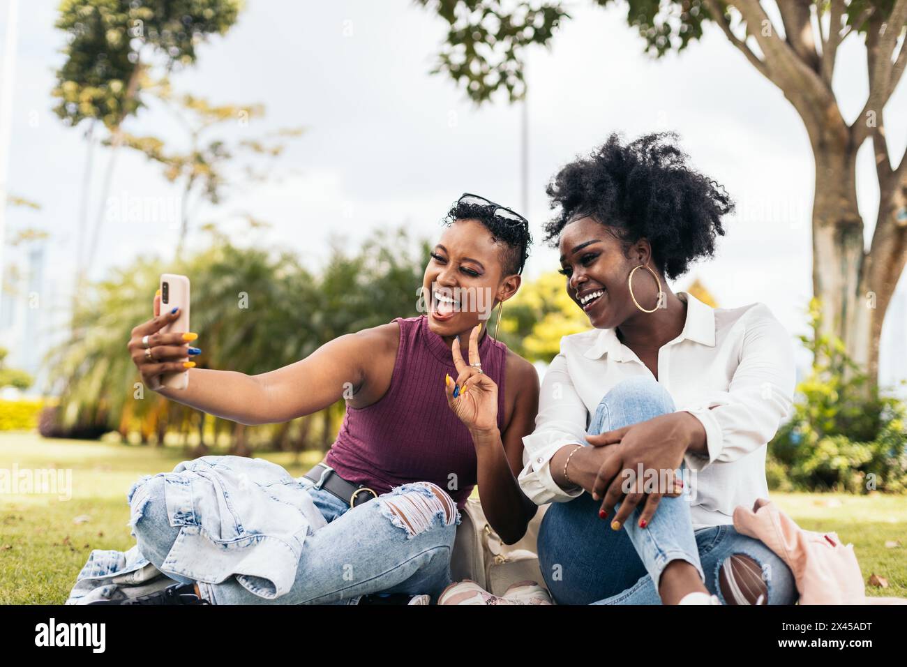 Deux amies noires s'amusant et prenant des selfies avec smartphone assis sur l'herbe dans le parc Banque D'Images