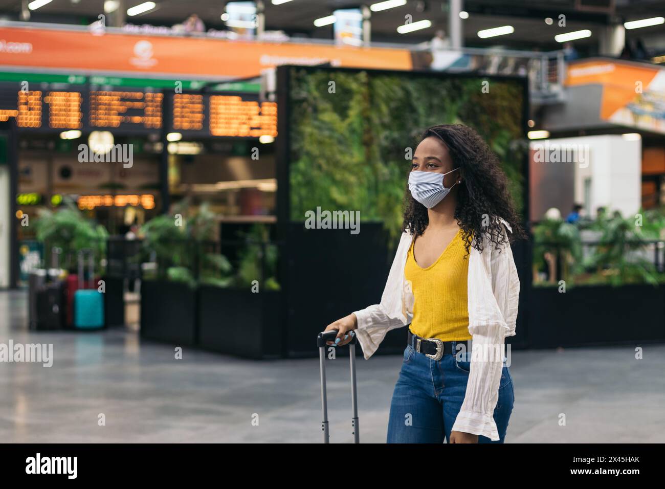 Jeune femme afro-américaine avec masque facial marchant sur la gare avec une valise après un voyage. Elle cherche son petit ami qui attend Banque D'Images