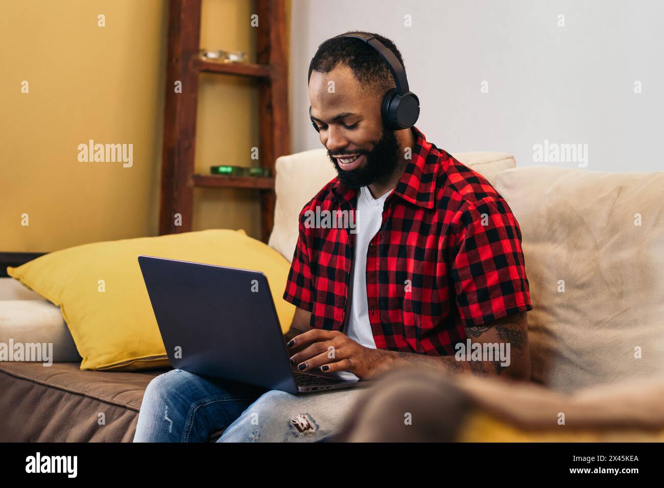 Homme noir souriant avec des ongles peints travaillant à distance de la maison avec un ordinateur portable et des écouteurs. Concept de télétravail et de bureau à domicile. Banque D'Images