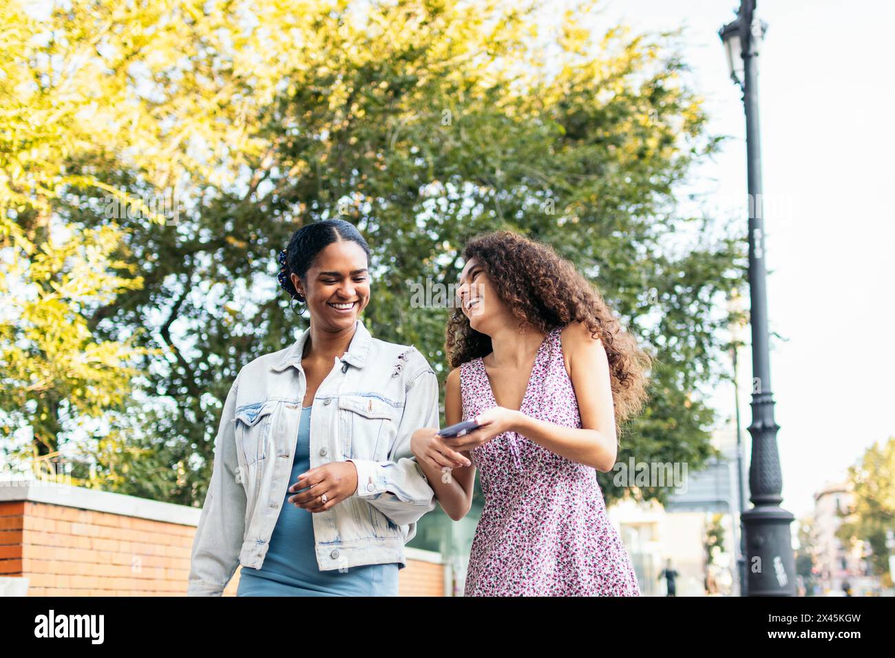 Deux amis latino-américains marchant dans la rue, s'amusant et riant en utilisant un smartphone. Banque D'Images