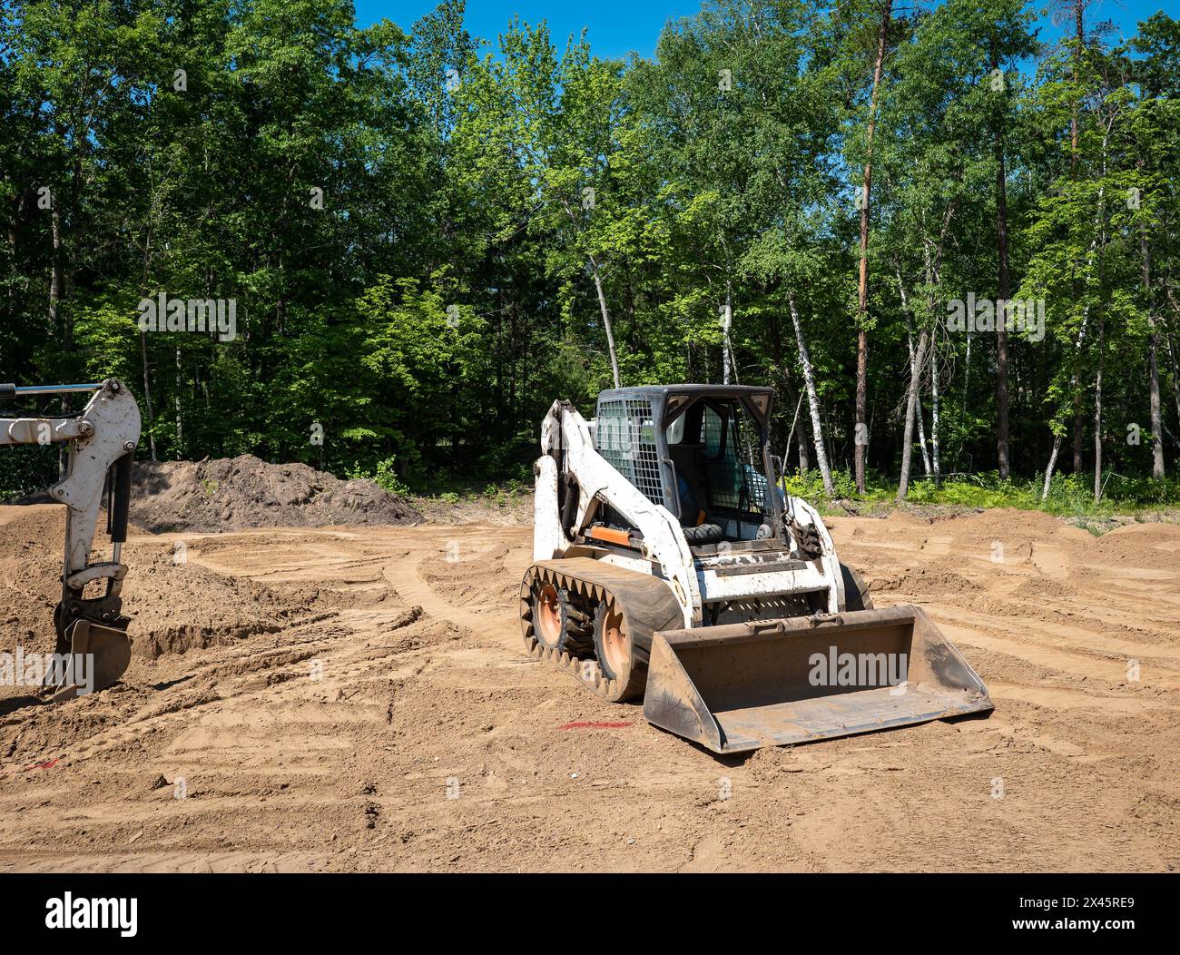 Chargeur frontal compact blanc, avec chenilles de mini-chargeuse, repose sur la terre dans un terrain vide bordé d'arbres, en préparation de la construction d'une nouvelle maison. Le Banque D'Images