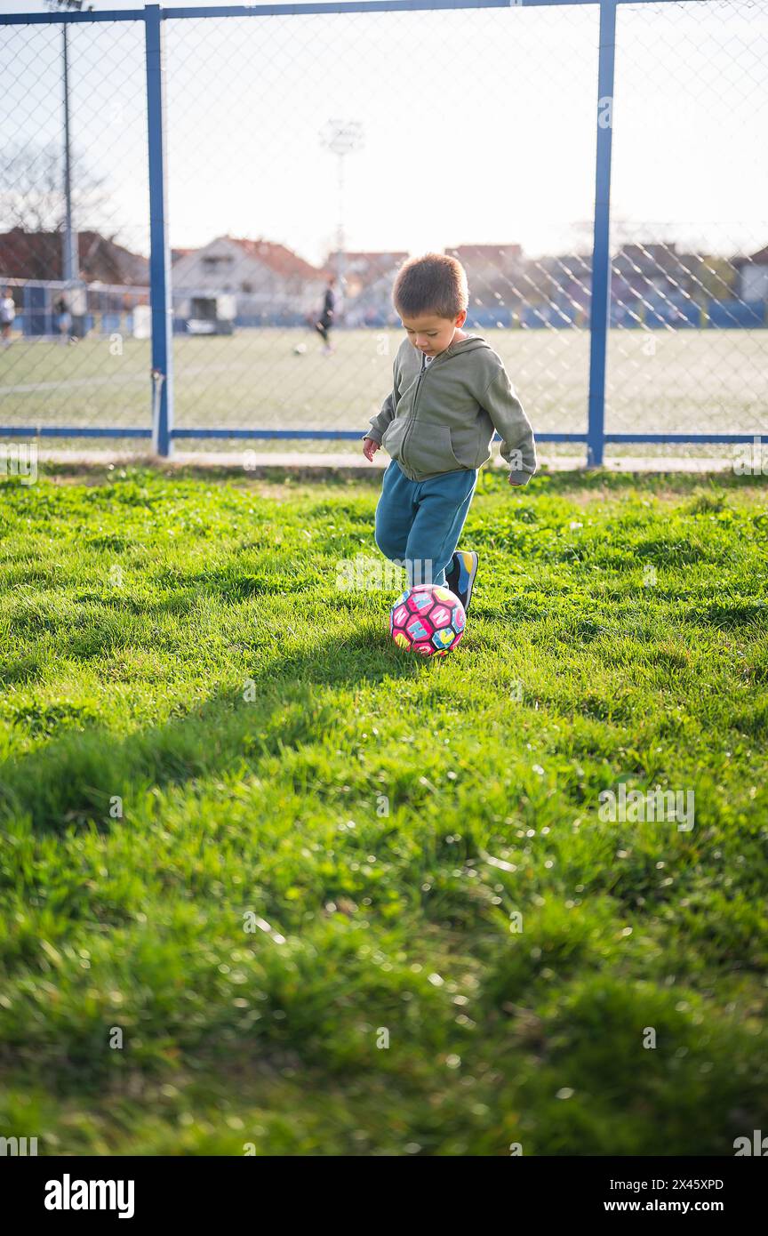 Capturé en plein match, ce garçon tout-petit se concentre sur le ballon de football, illustrant l'engagement d'un enfant de 3 ans avec le football sur herbe. Une scène vibrante capt Banque D'Images