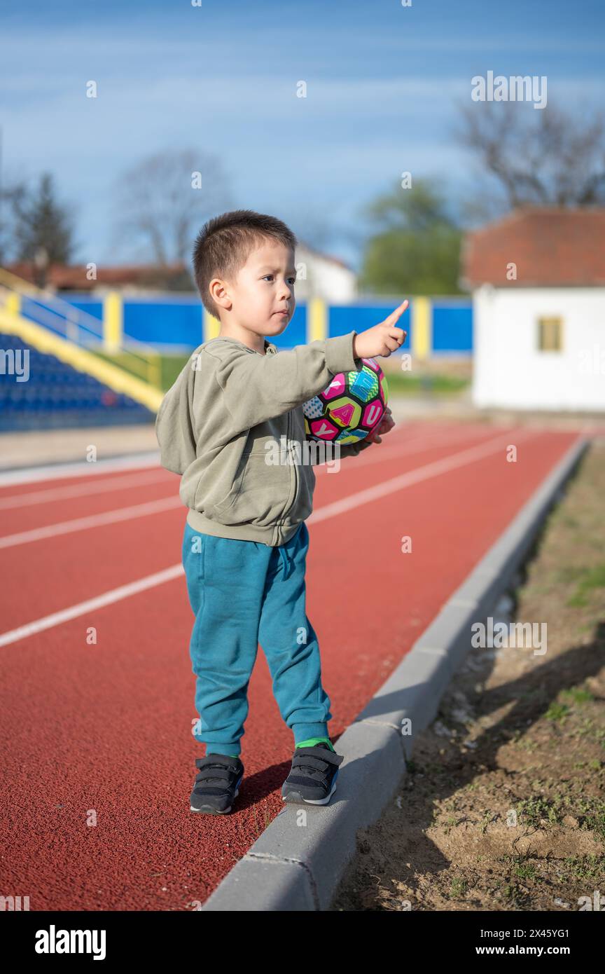 Un jeune garçon joyeux saisit un ballon de football dynamique sur la piste, signalant l'intérêt de l'enfant de 3 ans pour le football dès son plus jeune âge Banque D'Images