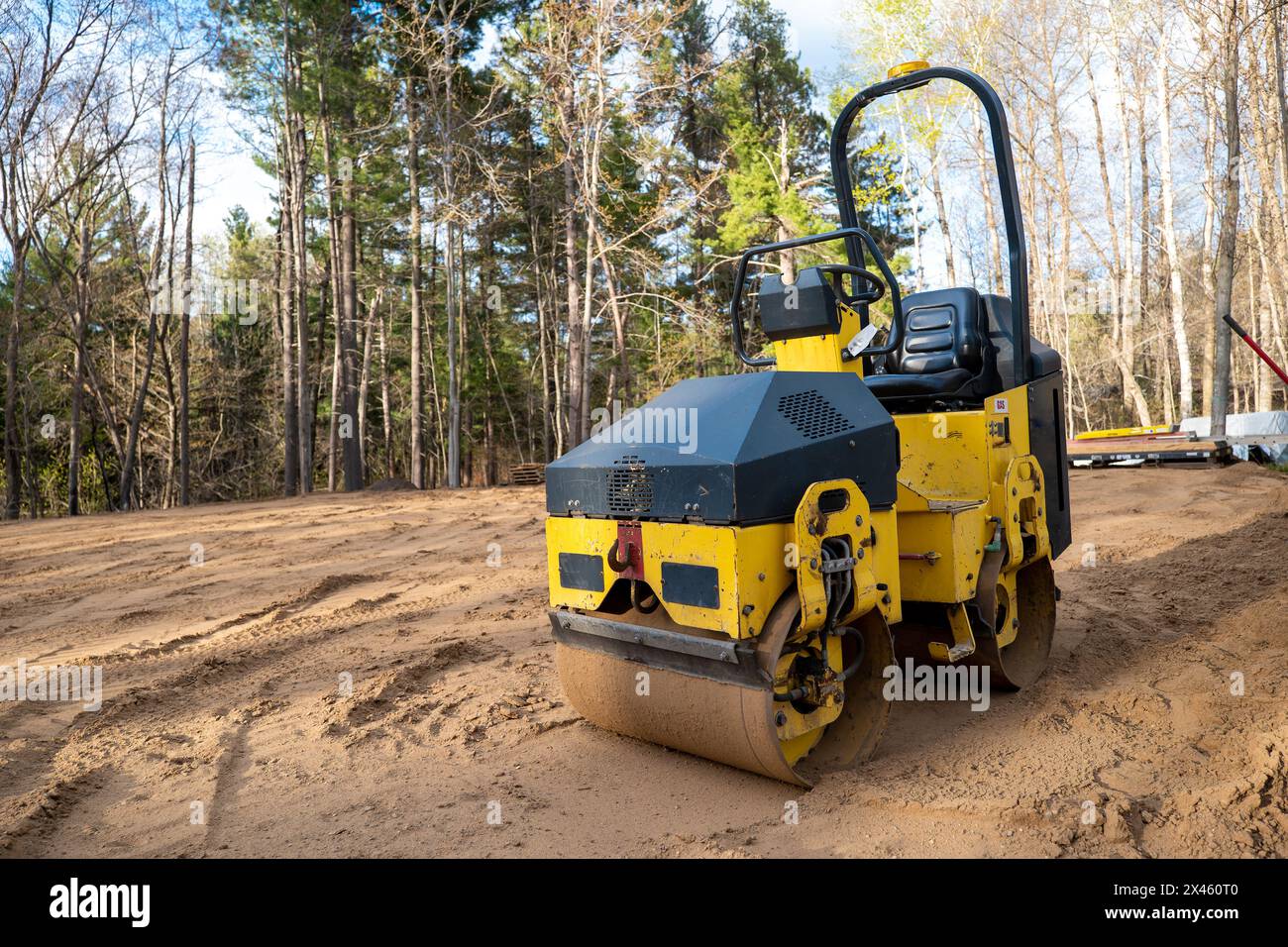Gros plan du rouleau de compacteur jaune, un usiné utilisé pour le compactage du sol, de la surface d'asphalte, du gravier et du sable. Vue sur un bâtiment vide bordé d'arbres Banque D'Images