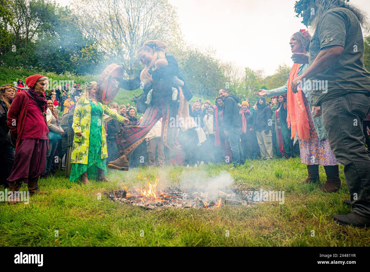 Les gens sautent le feu pendant les célébrations de Beltane à Glastonbury Chalice Well, où ils observent une interprétation moderne de l'ancien rite de fertilité païenne celtique du printemps. Date de la photo : mercredi 1er mai 2024. Banque D'Images