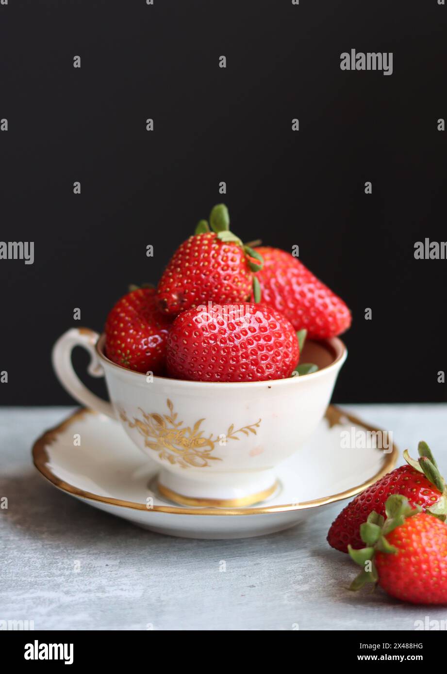 Fraises dans une petite tasse en porcelaine blanche et dorée sur fond sombre avec espace pour le texte. Photo de natures mortes avec vaisselle rétro et fruits d'été. E Banque D'Images