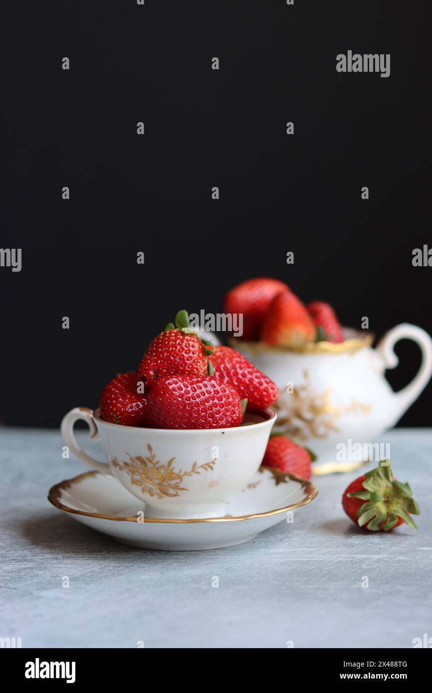 Fraises dans une petite tasse en porcelaine blanche et dorée sur fond sombre avec espace pour le texte. Photo de natures mortes avec vaisselle rétro et fruits d'été. E Banque D'Images