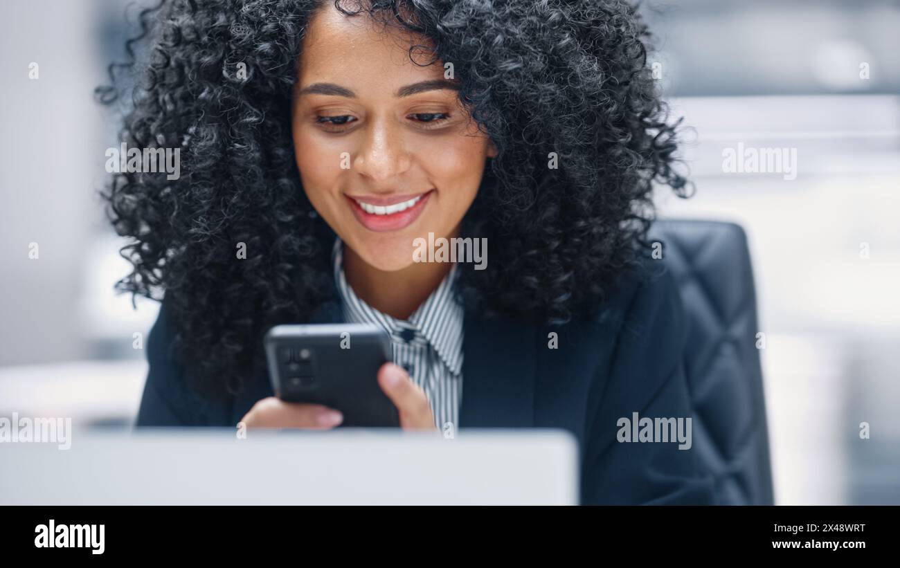 Degré moderne : femme d'affaires noire assise à son bureau à l'aide d'un smartphone. Entrepreneur numérique afro-américain utilisant le téléphone portable, Internet e-commerce Online Shopping. Prise de vue avec un arrière-plan flou de mouvement Banque D'Images