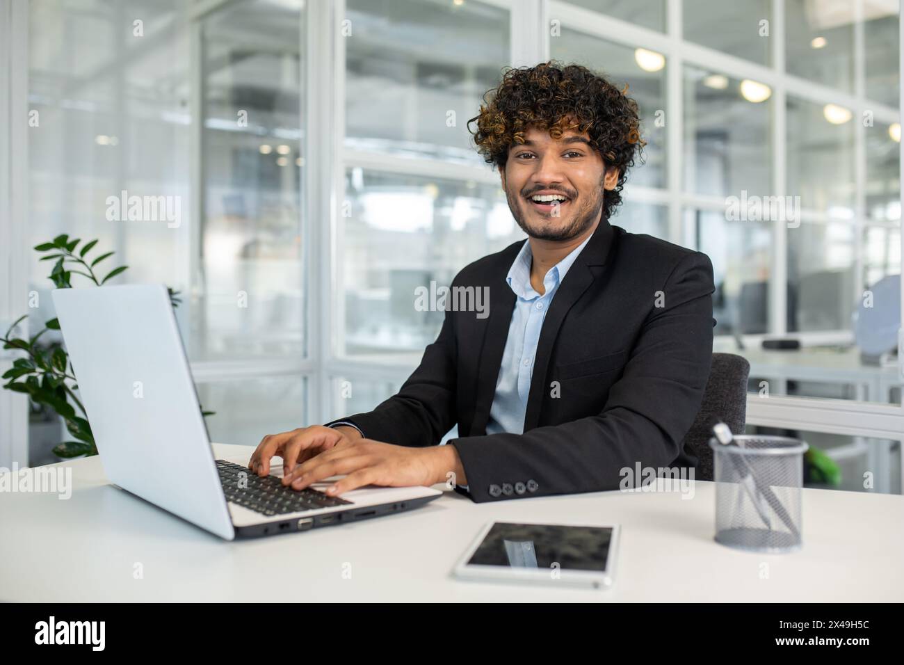 Homme d'affaires arabe gardant les mains sur le clavier de l'ordinateur portable tout en regardant la caméra avec le sourire dentelé. Employeur bouclé se sentant prêt à commencer une nouvelle semaine productive et rentable au bureau. Banque D'Images