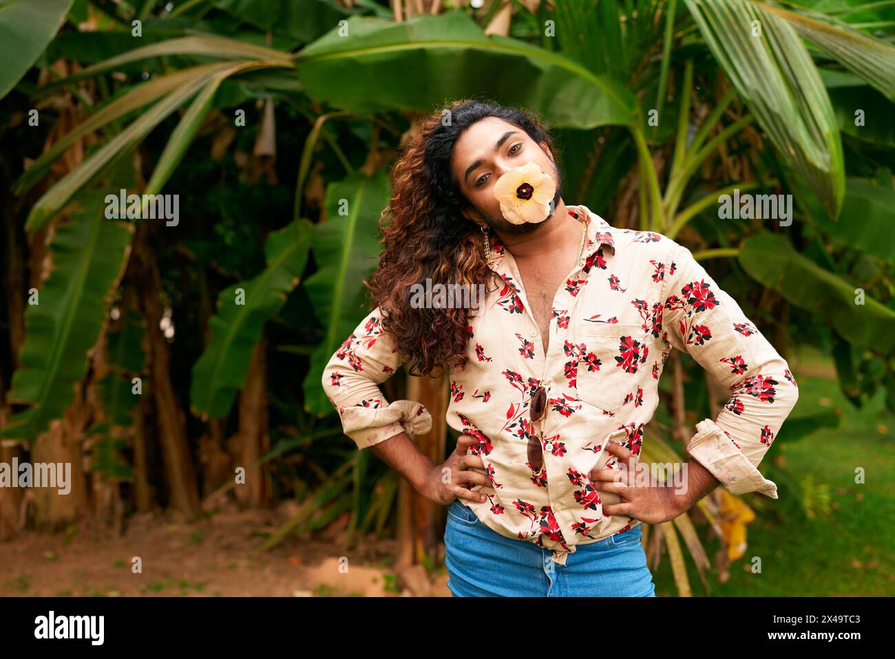 Homme noir confiant avec une fleur jaune dans la bouche pose dans la nature. Modèle masculin gay africain avec cheveux bouclés, chemise florale, poutres de jeans bleus Banque D'Images