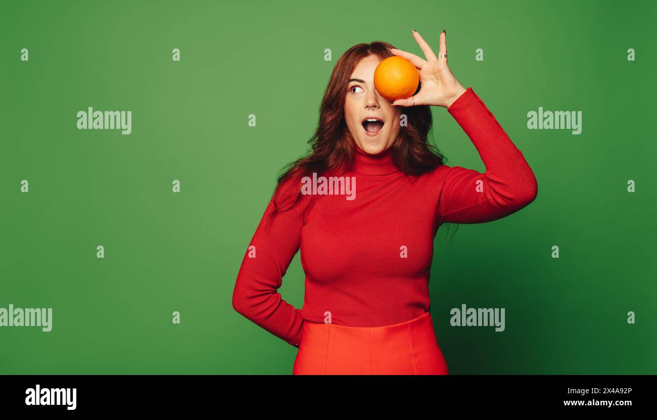 Femme joyeuse se tient sur un fond vert, tenant un agrume orange. Ses vêtements décontractés et son style tendance reflètent sa vie fraîche et saine Banque D'Images