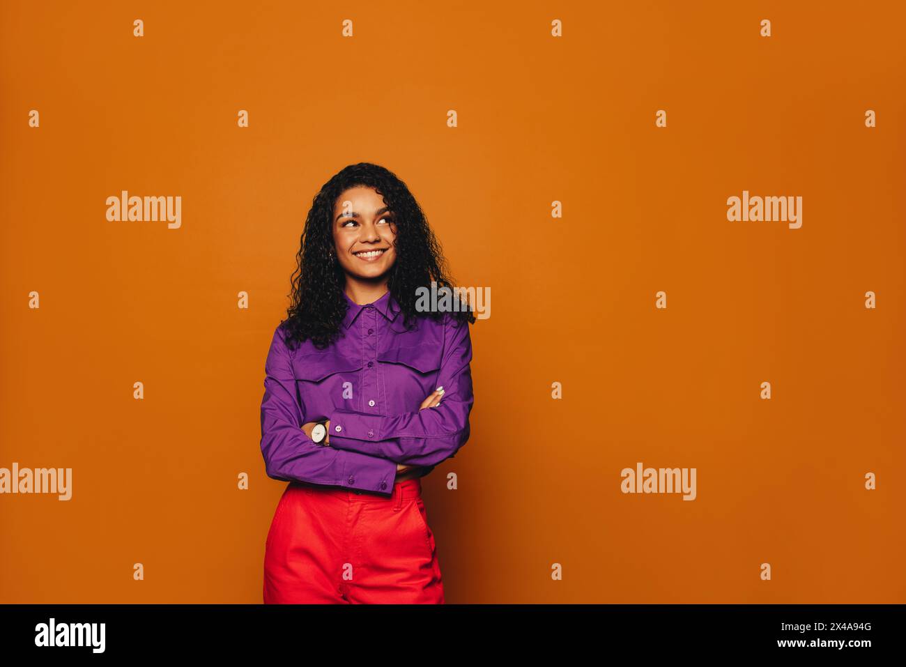 Femme heureuse avec les cheveux bouclés, dans des vêtements décontractés, se tient soigneusement dans un studio vibrant. Son sourire éclatant et son expression pensive révèlent son élégant A. Banque D'Images