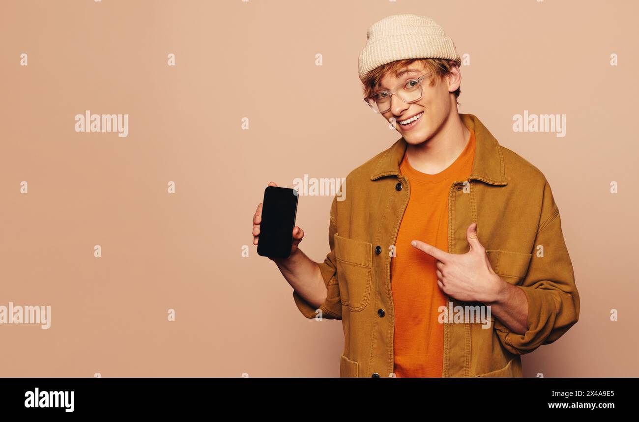 Jeune homme, en vêtements décontractés et veste en denim, tient un téléphone montrant une application mobile. Pointant vers l'écran tactile, il le recommande gaiement. La vibr Banque D'Images