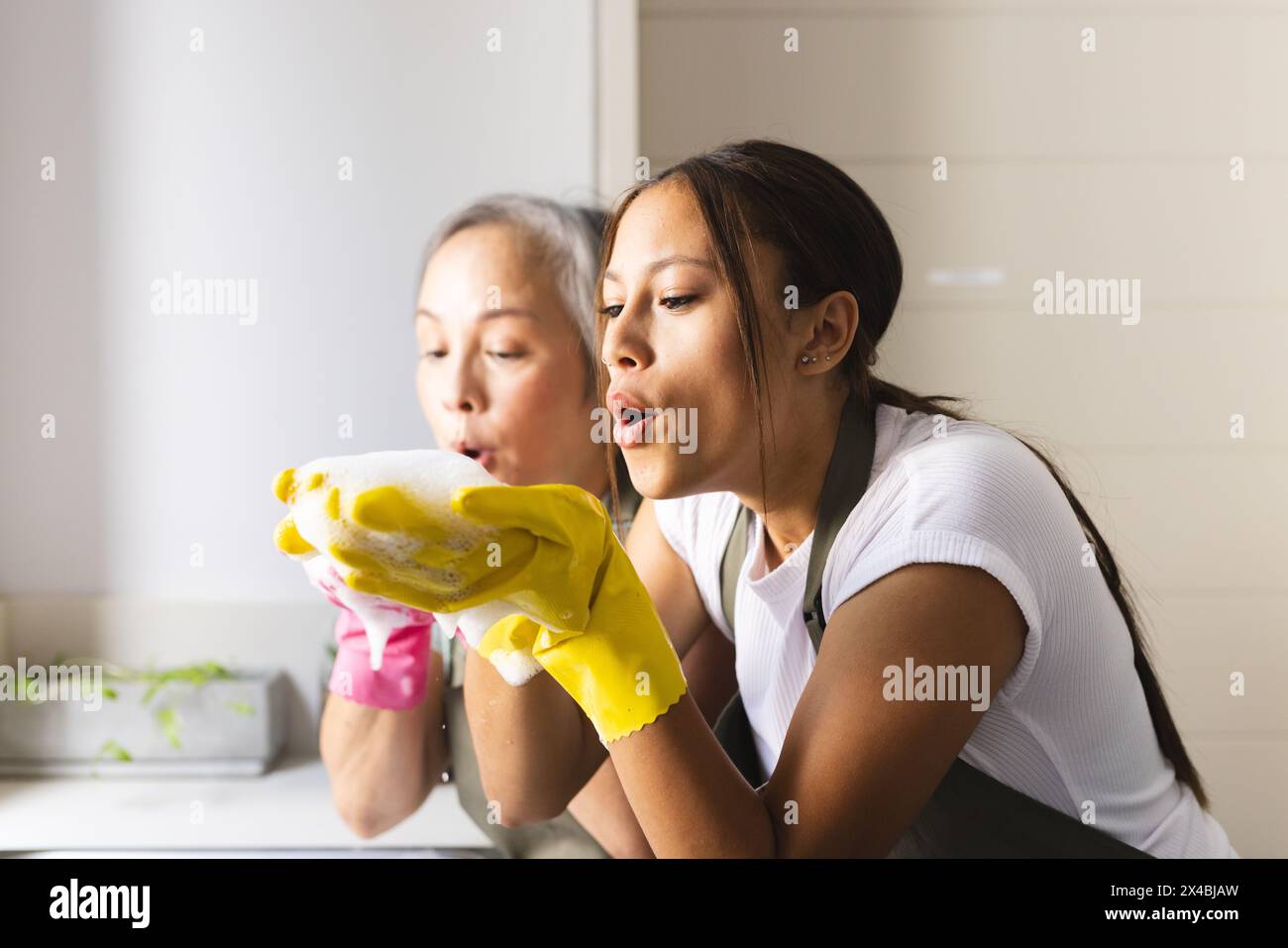 Grand-mère asiatique et petite-fille adolescente biraciale soufflant des bulles à la maison. Tous deux habillés de façon décontractée, profitant d'un moment ludique en cuisine, avec le gran Banque D'Images