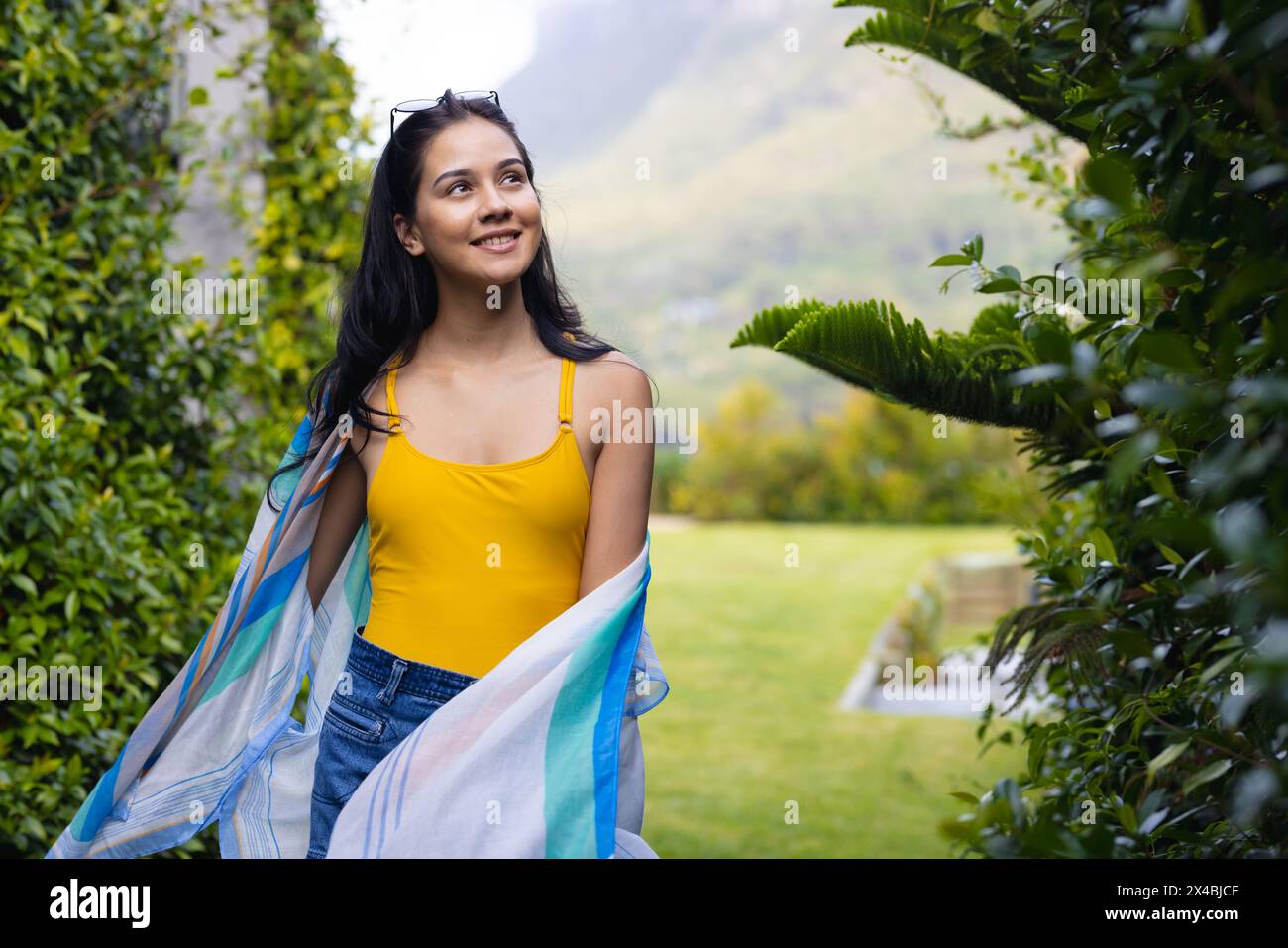 Jeune femme biraciale portant un haut jaune debout à l'extérieur à la maison. Arborant de longs cheveux foncés, souriant, regardant loin de la caméra, elle rayonne de chaleur et Banque D'Images Jeune femme biraciale portant un haut jaune debout à l'extérieur à la maison. Arborant de longs cheveux foncés, souriant, regardant loin de la caméra, elle rayonne de chaleur et Banque D'Images