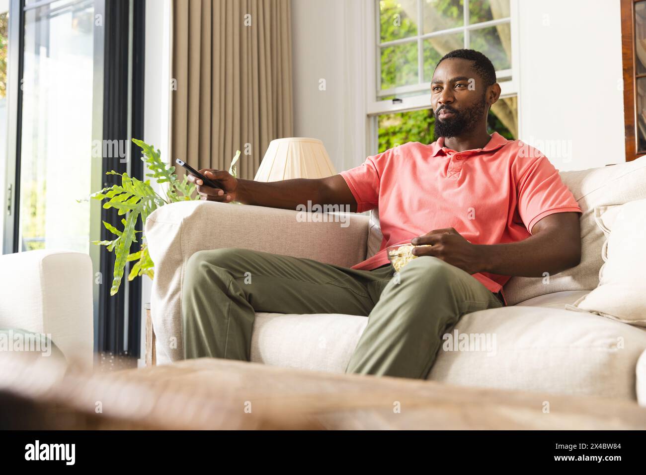 À la maison, homme afro-américain portant une chemise rose et un pantalon vert, tenant à distance. Arborant une barbe courte et dégageant une ambiance détendue, assis sur un sof Banque D'Images À la maison, homme afro-américain portant une chemise rose et un pantalon vert, tenant à distance. Arborant une barbe courte et dégageant une ambiance détendue, assis sur un sof Banque D'Images