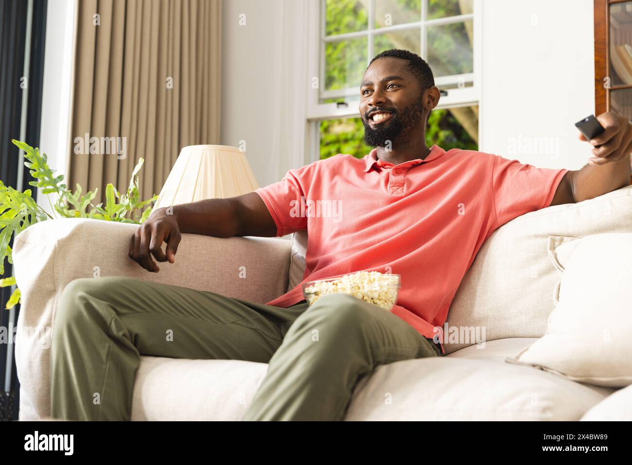Homme afro-américain se relaxant sur le canapé à la maison, tenant à distance. Il a les cheveux noirs courts, portant une chemise rouge décontractée et un pantalon vert, inaltérés Banque D'Images Homme afro-américain se relaxant sur le canapé à la maison, tenant à distance. Il a les cheveux noirs courts, portant une chemise rouge décontractée et un pantalon vert, inaltérés Banque D'Images