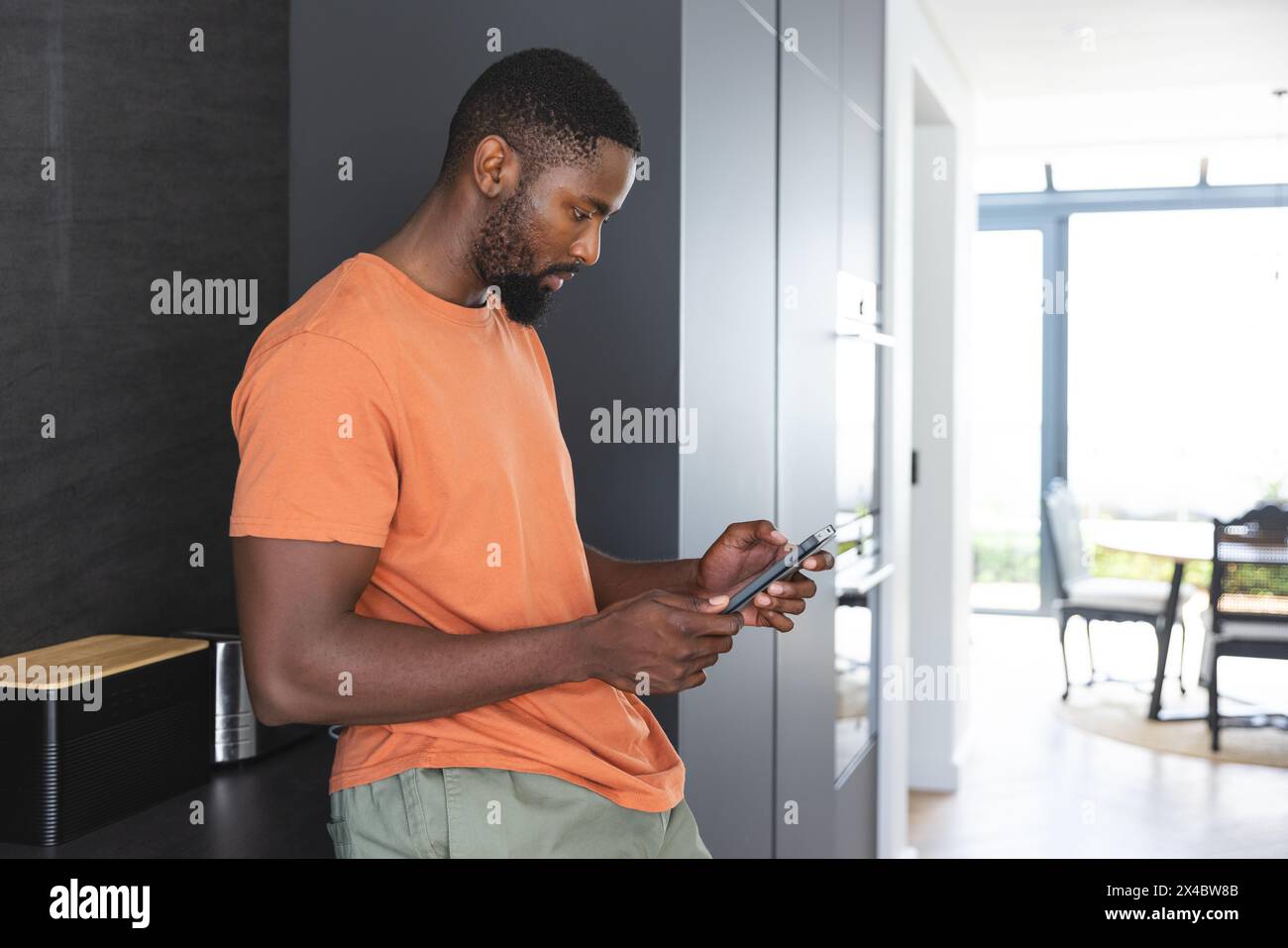 À la maison, un homme afro-américain vérifiant smartphone, portant une chemise orange. Sport cheveux courts, profiter d'un moment calme dans la cuisine bien éclairée avec un vert Banque D'Images À la maison, un homme afro-américain vérifiant smartphone, portant une chemise orange. Sport cheveux courts, profiter d'un moment calme dans la cuisine bien éclairée avec un vert Banque D'Images