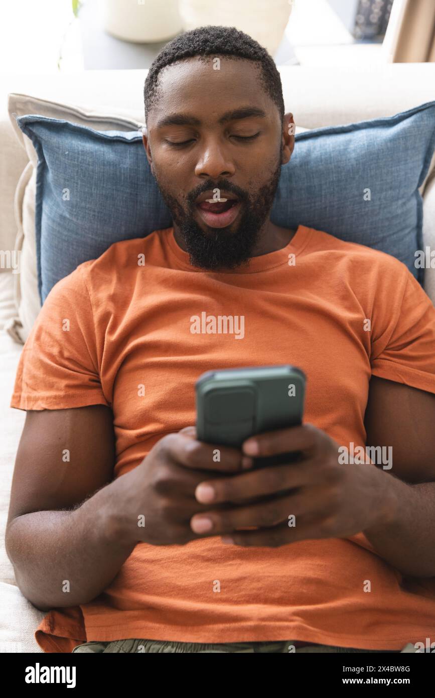Afro-américain propriétaire assis, axé sur le smartphone. Il a les cheveux noirs courts, la barbe, portant une chemise orange, inaltérée Banque D'Images