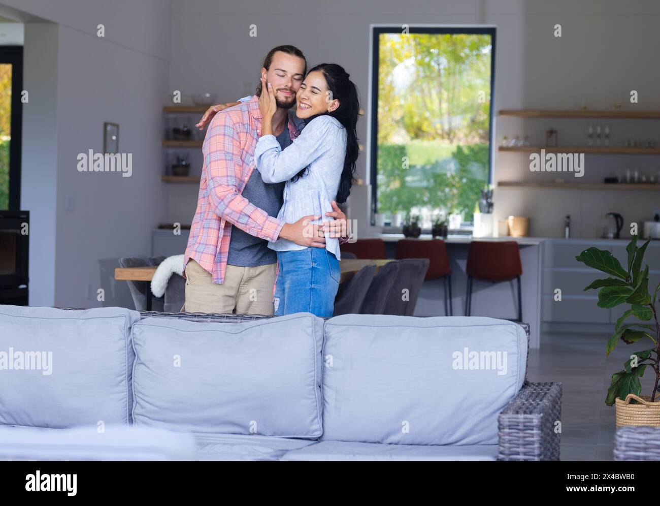 Couple diversifié embrassant dans un salon lumineux à la maison, espace de copie. Elle a les cheveux foncés et il porte une barbe, tous deux portant des vêtements décontractés, inaltérés Banque D'Images Couple diversifié embrassant dans un salon lumineux à la maison, espace de copie. Elle a les cheveux foncés et il porte une barbe, tous deux portant des vêtements décontractés, inaltérés Banque D'Images