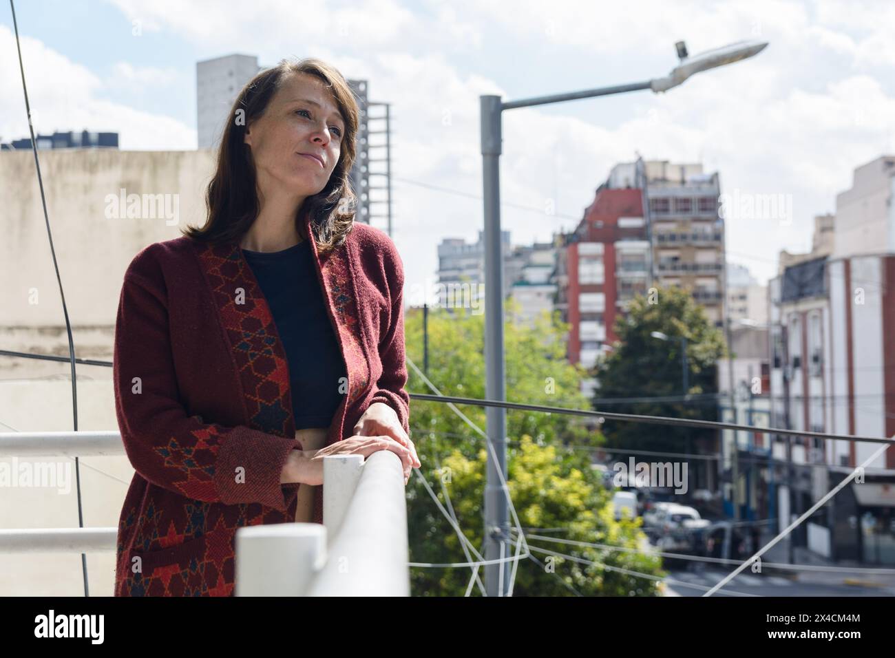 Femme adulte latine avec veste pour le froid est en amour, debout sur le balcon de son appartement seul, regardant devant, réfléchie, contemplant avec le visage triste, Banque D'Images