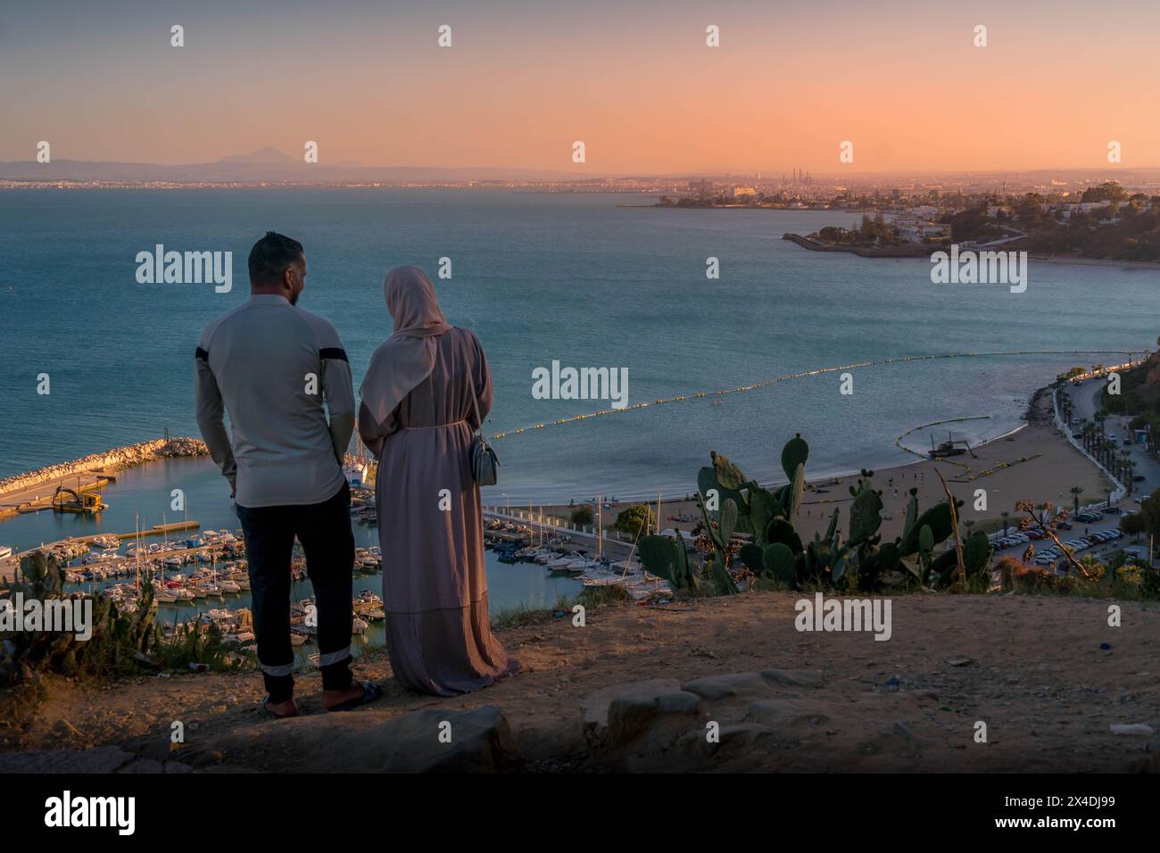Le couple debout sur le bord de la falaise à Sidi Bou Said, regardant vers le bas sur la mer Méditerranée, le panorama de la ville de Tunis et le coucher de soleil pittoresque. Banque D'Images