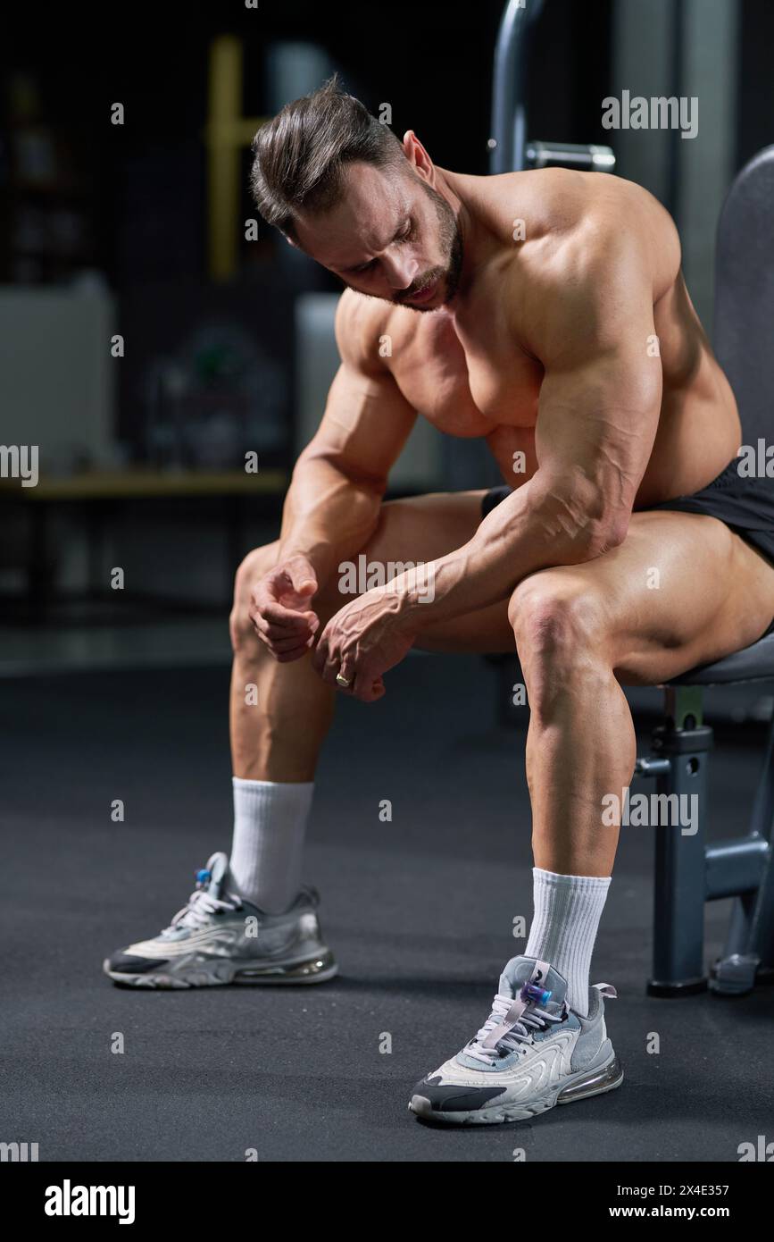 Bodybuilder caucasien sérieux ayant le repos après l'exercice dans le gymnase. Vue latérale de l'athlète masculin fatigué avec un corps fort assis sur la chaise, appuyé sur les jambes, Banque D'Images Bodybuilder caucasien sérieux ayant le repos après l'exercice dans le gymnase. Vue latérale de l'athlète masculin fatigué avec un corps fort assis sur la chaise, appuyé sur les jambes, Banque D'Images