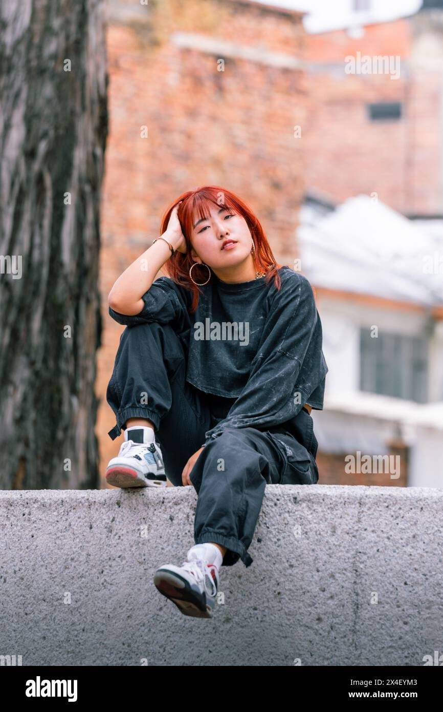 Femme coréenne réfléchie avec des cheveux roux vibrants assis décontractés dans un lieu urbain Banque D'Images