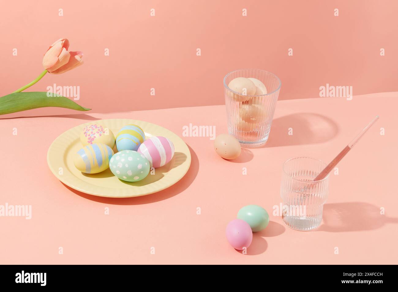 Oeufs de Pâques colorés décorés sur un plat jaune avec des tasses de fleurs et d'eau sur un fond rose pastel. Les œufs symbolisent la fertilité à Pâques. Main spéciale Banque D'Images